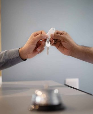 Unrecognizable Hotel Receptionist Giving Key To Male Guest At Reception In Lobby, Closeup Of Hands. Tourist Checking In At Accomodation, Taking Room Keys From Hostel Worker. Cropped Shot